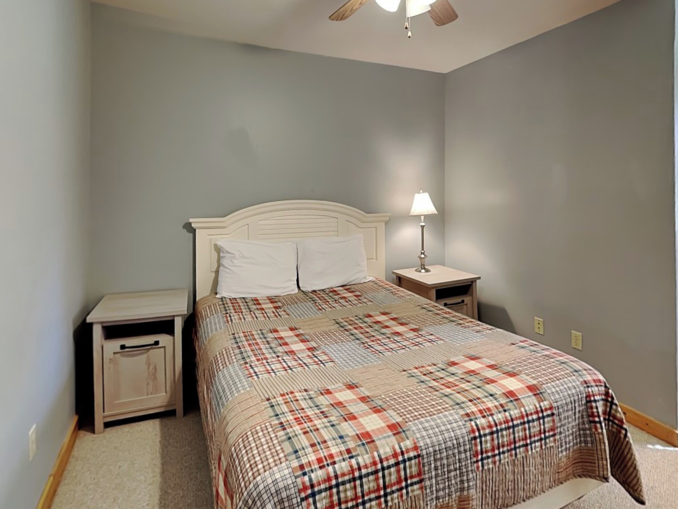 Une chambre à coucher comprenant un lit, deux tables de chevet, une commode, un ventilateur de plafond et une prise électrique murale.