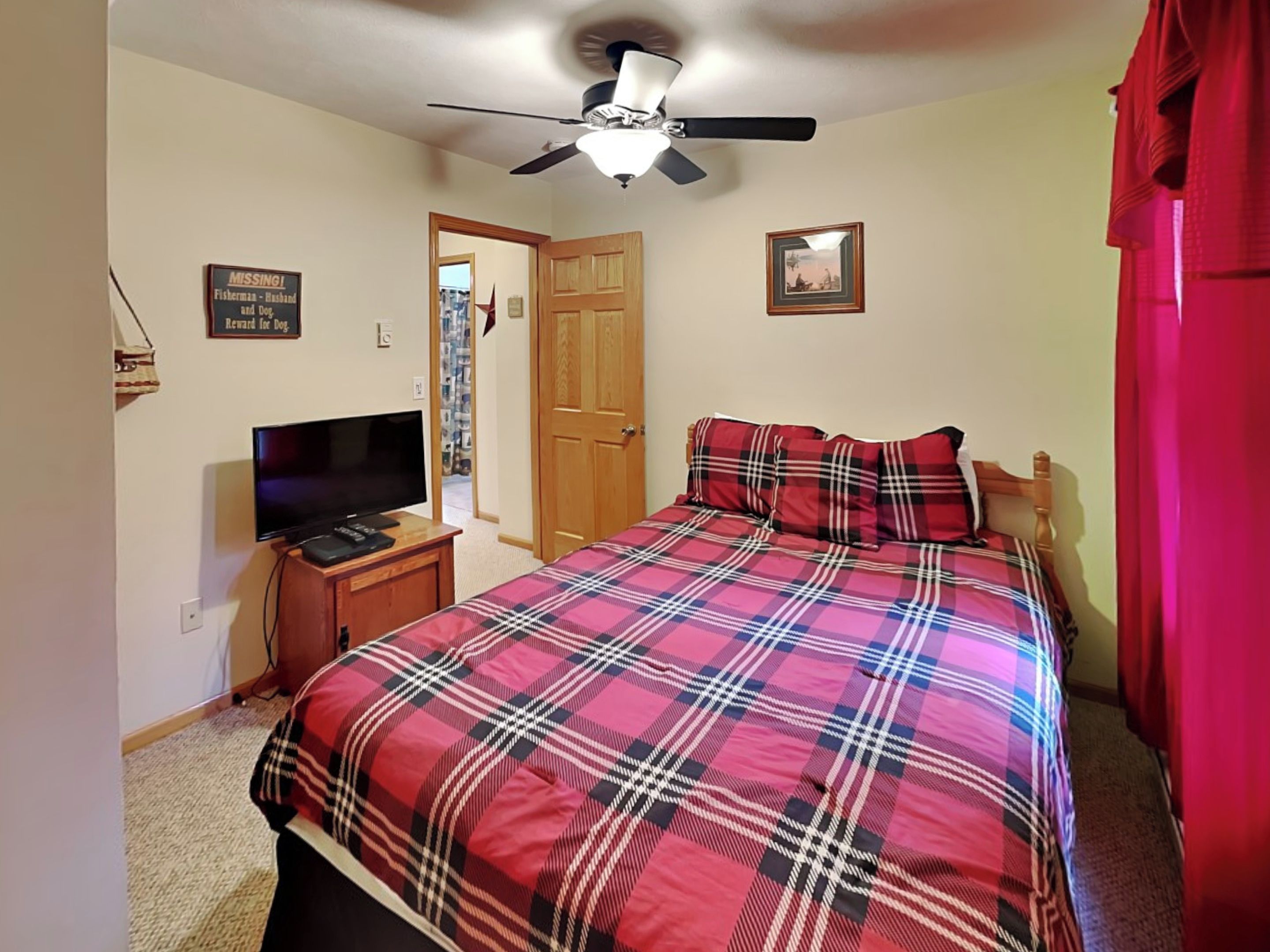 A bedroom with a bed, a television on a stand, a ceiling fan, and a door leading to another room.
