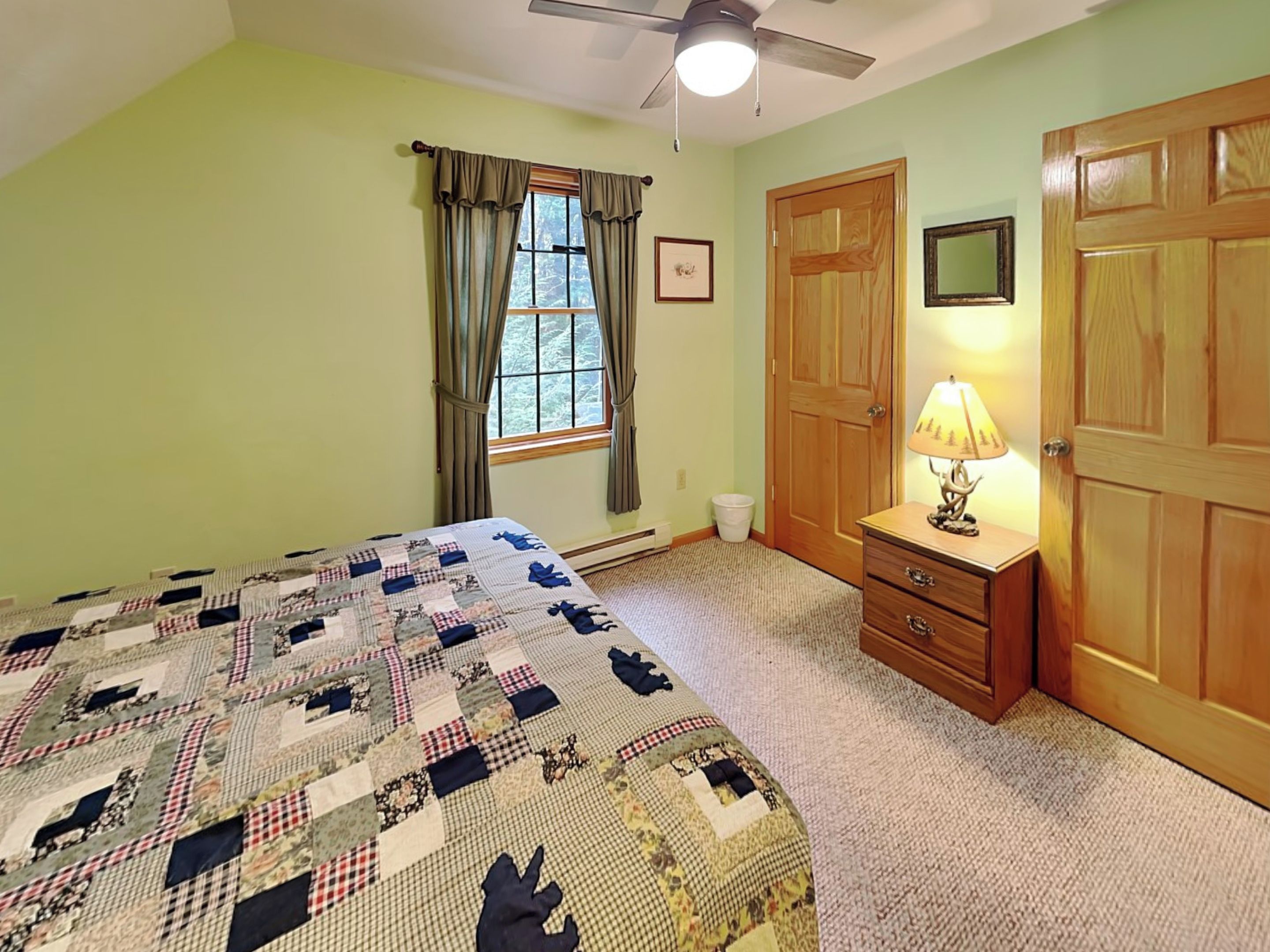Un dormitorio con cama, mesita de noche, lámpara, ventilador de techo, ventana con cortinas y puerta.