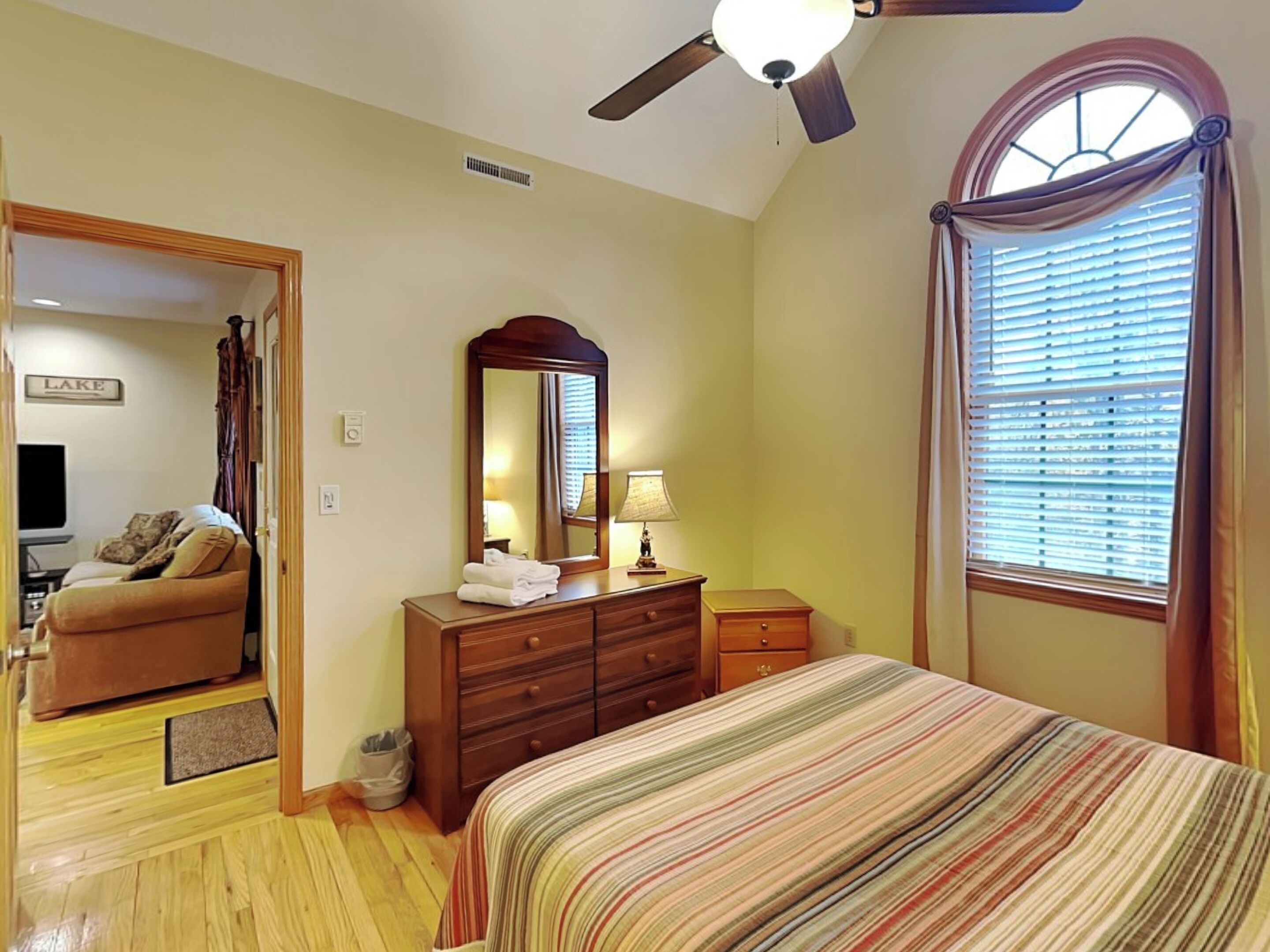 A bedroom with a bed, a dresser, a mirror, a ceiling fan, and a window with blinds.