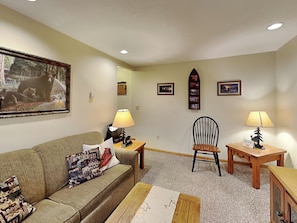 A living room with a sofa, a coffee table, a wooden side table, a chair, and a lamp. There are framed pictures on the wall.