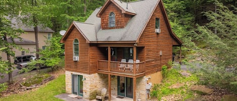 A two-story log cabin with a stone foundation, a porch with outdoor seating, and a forested backdrop.