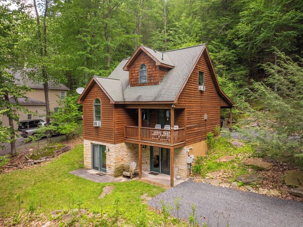 A two-story log cabin with a stone foundation, a porch with outdoor seating, and a forested backdrop.