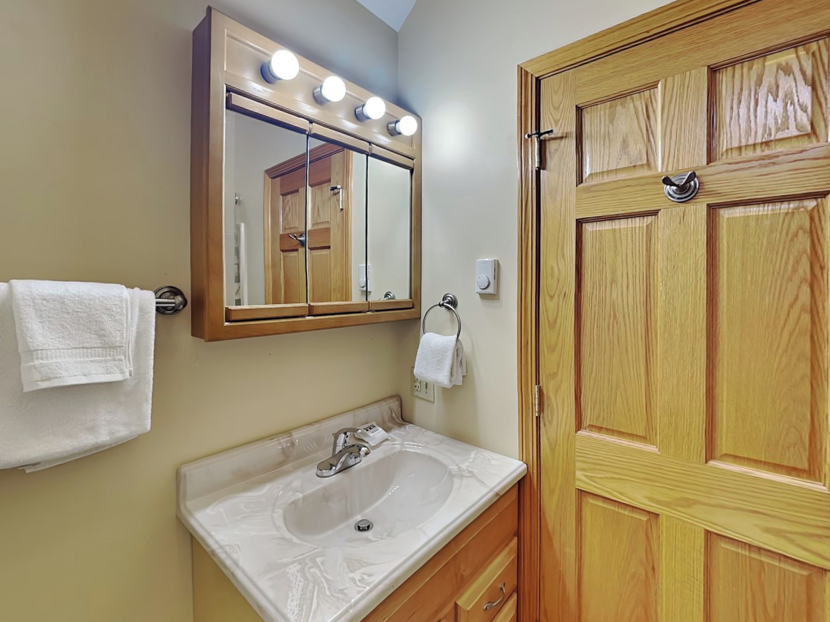 A bathroom with a wooden door, a mirror with lights, a sink, and two towels.