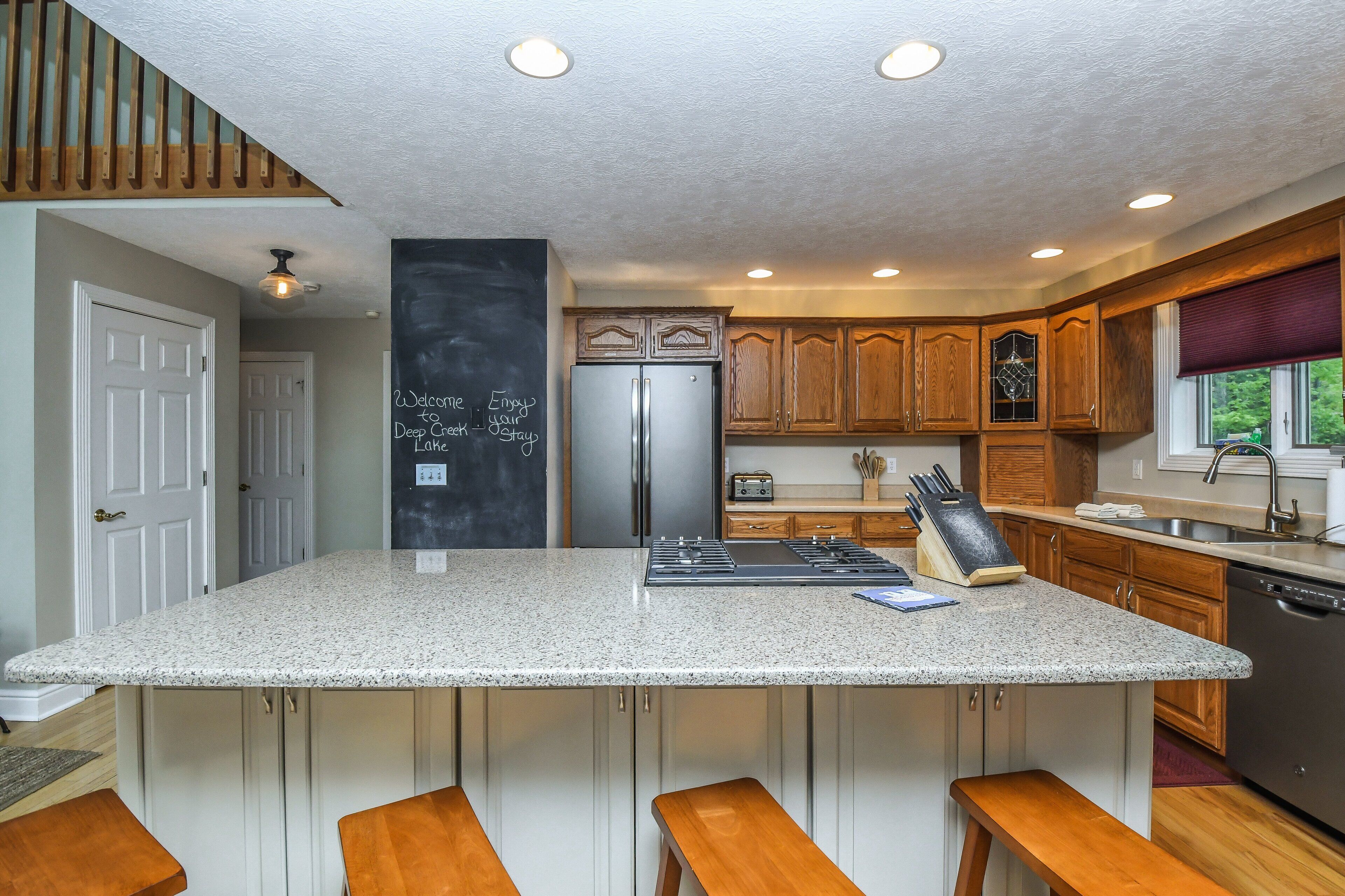 A kitchen with a large island, wooden cabinets, a stainless steel refrigerator, and a chalkboard with a welcome message.