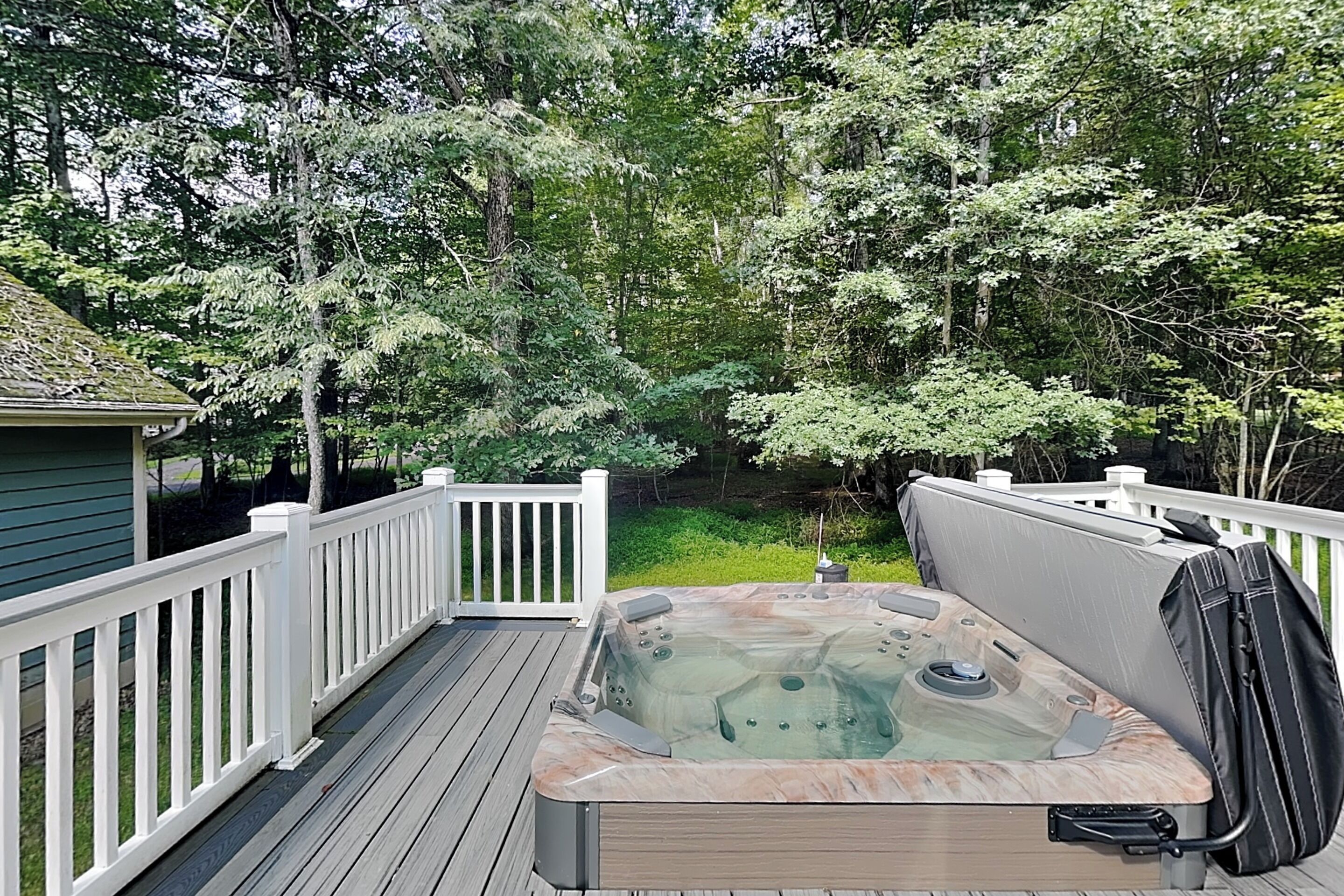 A hot tub on a wooden deck with white railings, surrounded by lush greenery.