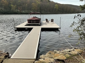 Un muelle de madera que se adentra en un lago, con un bote cubierto y dos sillas sobre una plataforma.