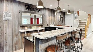 A kitchen with a marble island, bar stools, pendant lights, and a wooden accent wall with a mirror and signs. - Hiatus by Taylor Made Deep Creek (McHenry)