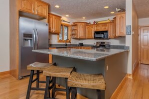 A kitchen with wooden cabinets, a stainless steel refrigerator, a granite countertop, and two bar stools.