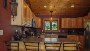 A kitchen with wooden cabinets, a central island with bar stools, a window with a view, and a hanging light fixture. - High-skis' Villa by Taylor Made Deep Creek (McHenry)