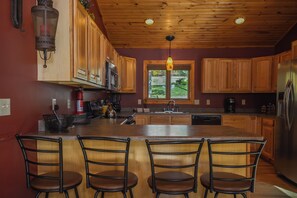 A kitchen with wooden cabinets, a central island with bar stools, a window with a view, and a hanging light fixture. - High-skis' Villa by Taylor Made Deep Creek (McHenry)