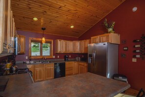 A kitchen with wooden cabinets, a stainless steel refrigerator, and a window with a view of greenery. - High-skis' Villa by Taylor Made Deep Creek (McHenry)