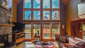 A spacious living room with a fireplace, leather sofa, and large windows offering a view of trees. - High-skis' Villa by Taylor Made Deep Creek (McHenry)