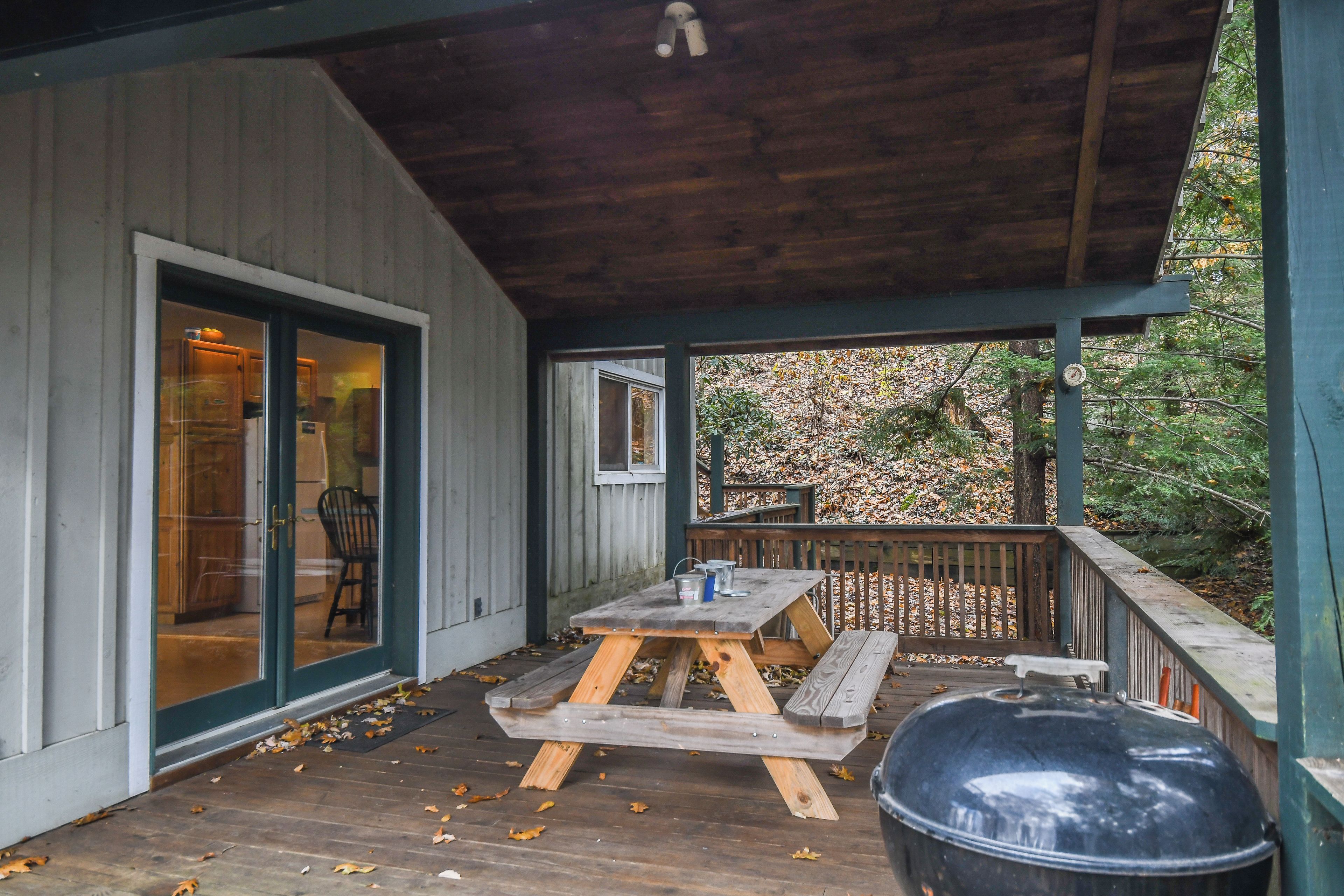 A covered patio with a wooden table and bench, a barbecue grill, and a view of a forested area.
