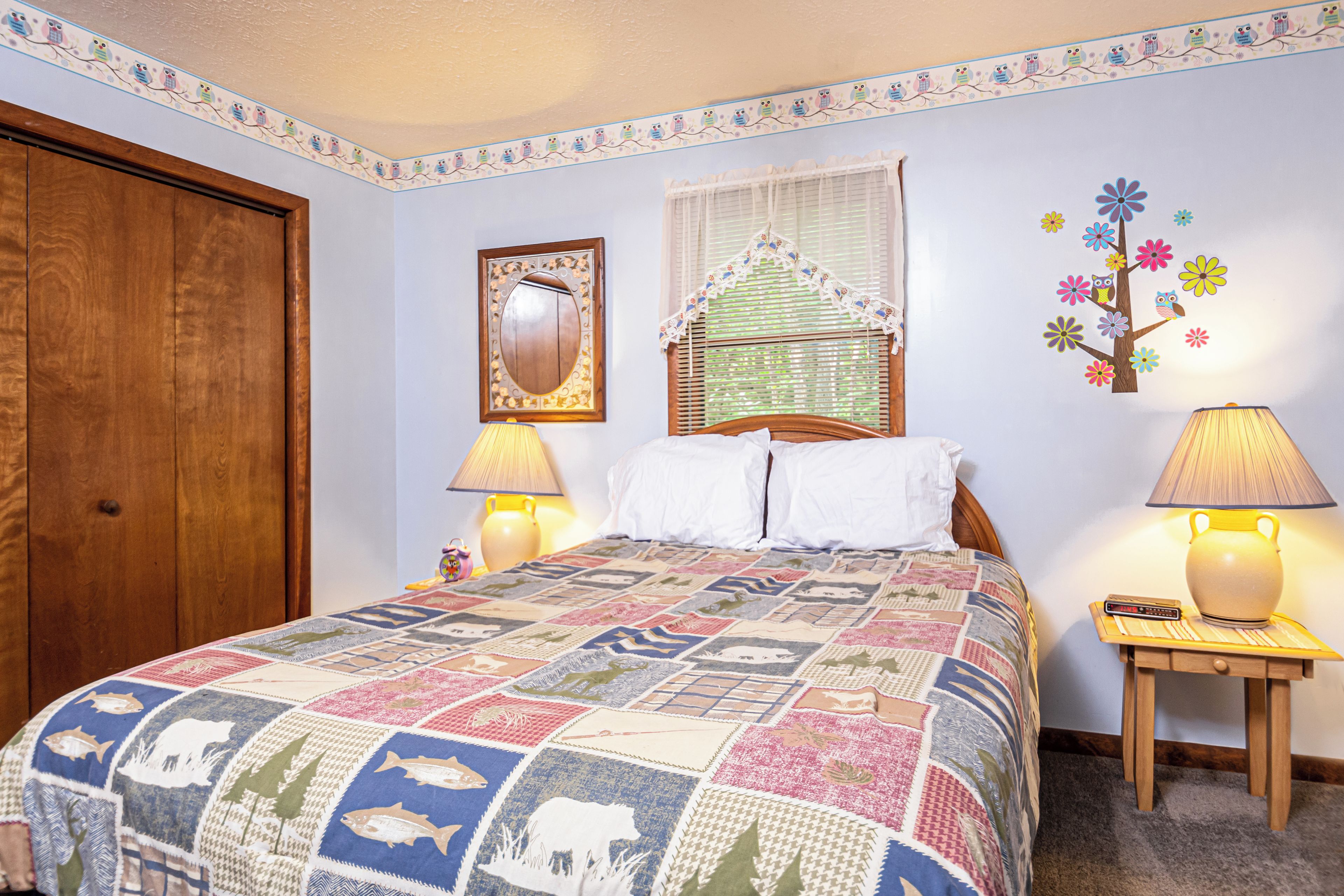 Un dormitorio con puerta de madera, una cama con colcha colorida, dos lámparas de noche, un espejo, una ventana con cortina de encaje y decoración de pared con un árbol y flores.