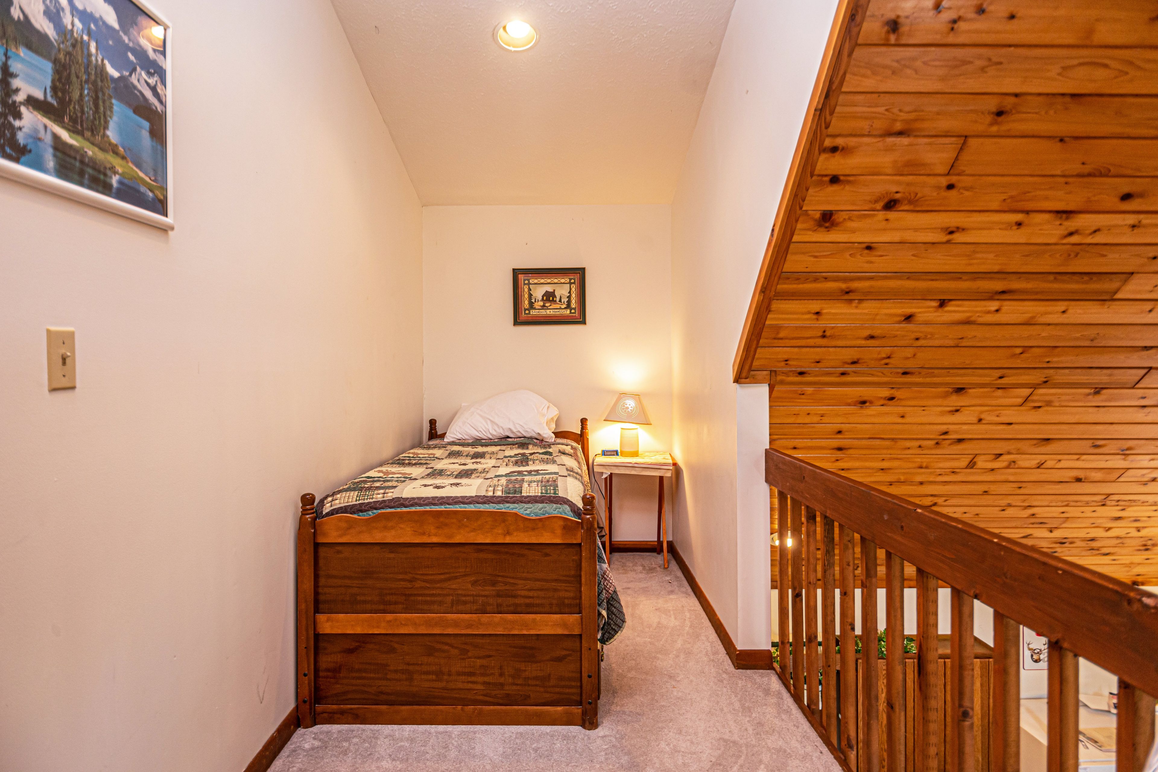 A bedroom with a wooden bed, a nightstand with a lamp, a framed picture on the wall, and a wooden staircase railing.