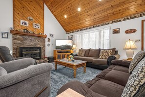 A cozy living room with a stone fireplace, wooden ceiling, and a large TV.