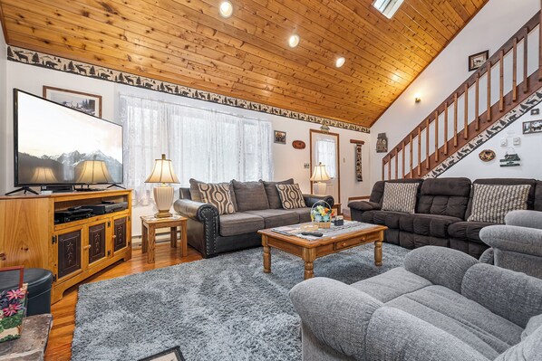 A cozy living room with wooden ceilings, a staircase, and a flat-screen TV.