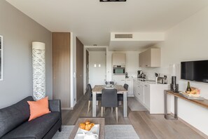 A modern living room with a sofa, coffee table, and a dining area with a table and chairs. A kitchen with a microwave and cabinets is visible in the background.