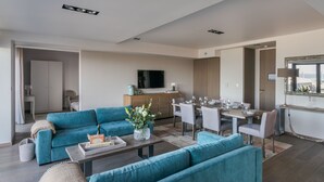 A modern living room with a teal sofa set, a dining area with a wooden table and chairs, and a kitchen area with cabinets and a TV.