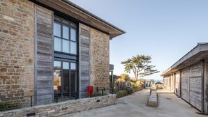 A modern building with stone walls and large windows, a concrete pathway, and a view of the ocean.