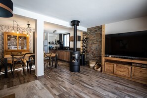 Uma sala de estar moderna com uma televisão grande, lareira, móveis de madeira e uma cozinha ao fundo.