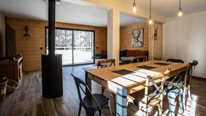 A modern interior with a large wooden dining table, black chairs, a central black stove, and a large window with a view of trees.