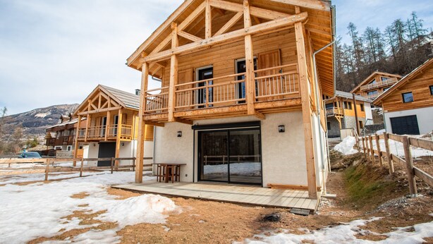 A wooden cabin with a balcony, a sliding glass door, and a view of the surrounding mountains.
