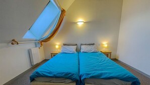 Een kamer met twee bedden, elk met een blauwe spreideken en witte kussens. Er staan nachtlampjes op houten nachtkastjes, een plafondlamp en een raam met gordijnen.