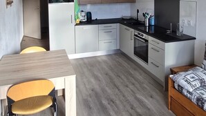 Een compacte keuken met witte kasten, een zwart aanrecht en een ingebouwde oven. Er is een houten eettafel met twee stoelen en een klein houten bankje.