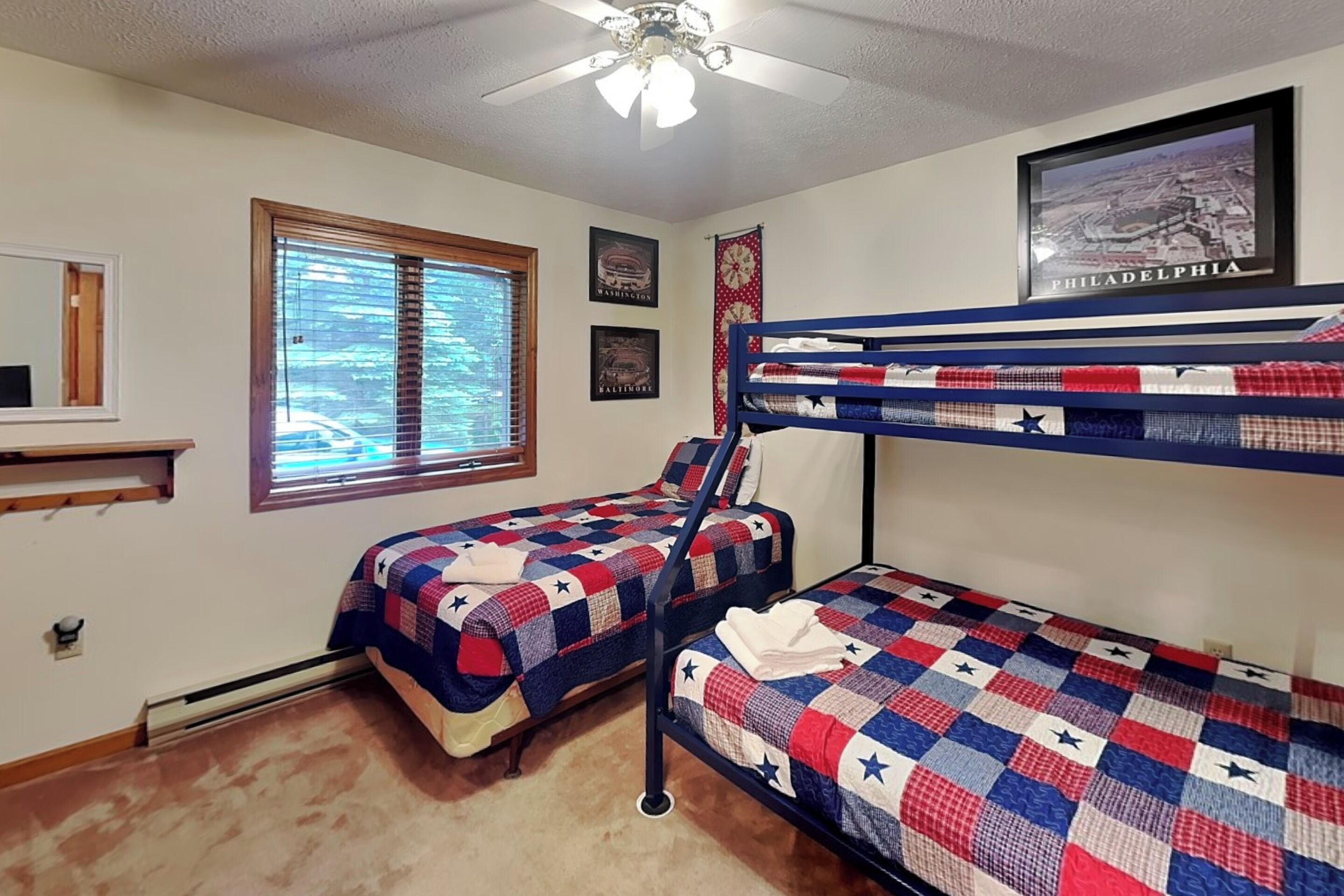 A room with bunk beds, a ceiling fan, a window with blinds, and a framed cityscape picture.