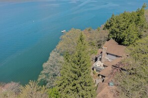 A house with a stone chimney, surrounded by dense trees, overlooking a calm lake with a boat. - Inspiration Point by Taylor Made Deep Creek (Swanton)