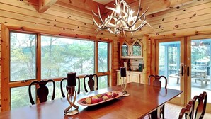 A wooden cabin interior with a dining table, chairs, a chandelier, and large windows offering a view of the outdoors. - Inspiration Point by Taylor Made Deep Creek (Swanton)