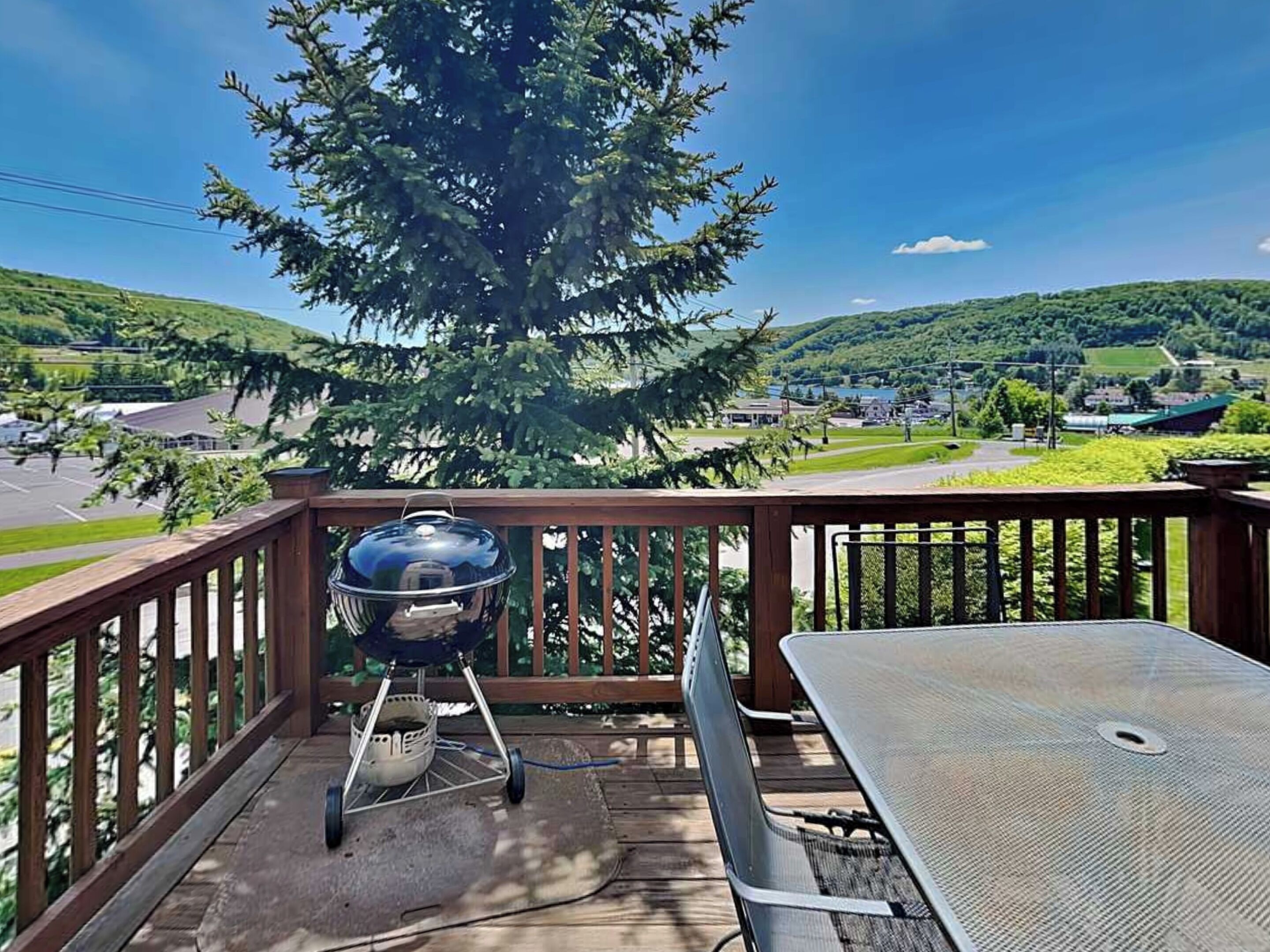 Een houten terras met een barbecue en een tafel, met uitzicht op een pittoresk landschap met heuvels en een meer.