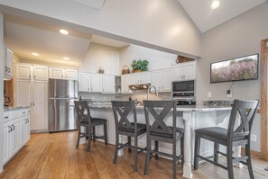 A modern kitchen with white cabinetry, a stainless steel refrigerator, a built-in oven, and a large island with bar stools. - Johnny Appleseed by Taylor Made Deep Creek (Oakland)