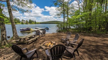 A lakeside campsite with a fire pit, picnic table, and Adirondack chairs.