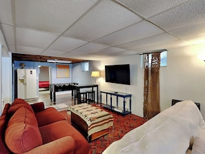 A living room with a sofa, ottoman, television, and a foosball table. - Just Around the Dilly by Taylor Made Deep Creek (Swanton)