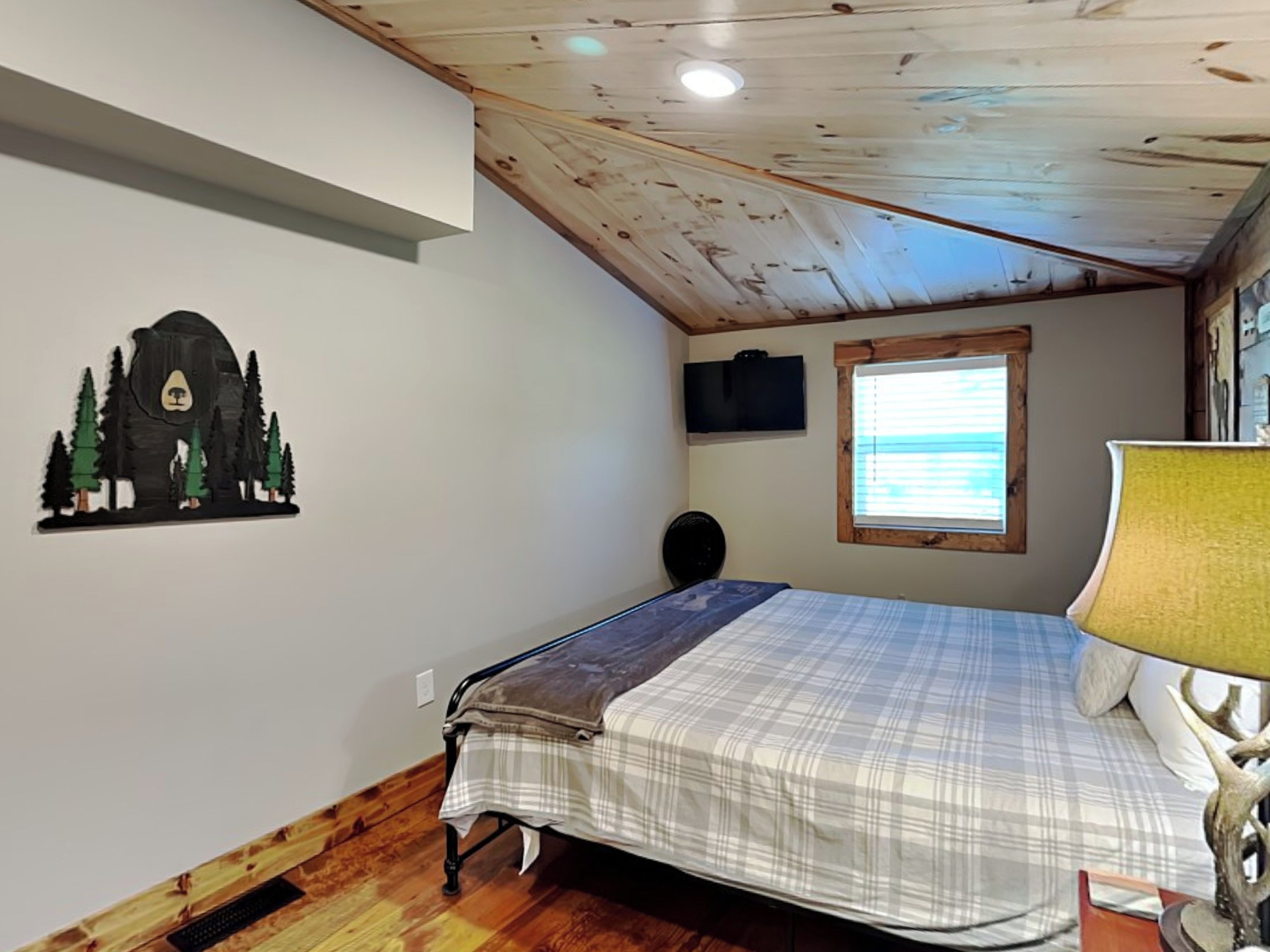 Un dormitorio con cama, ventana, televisor instalado en la pared y techo de madera.
