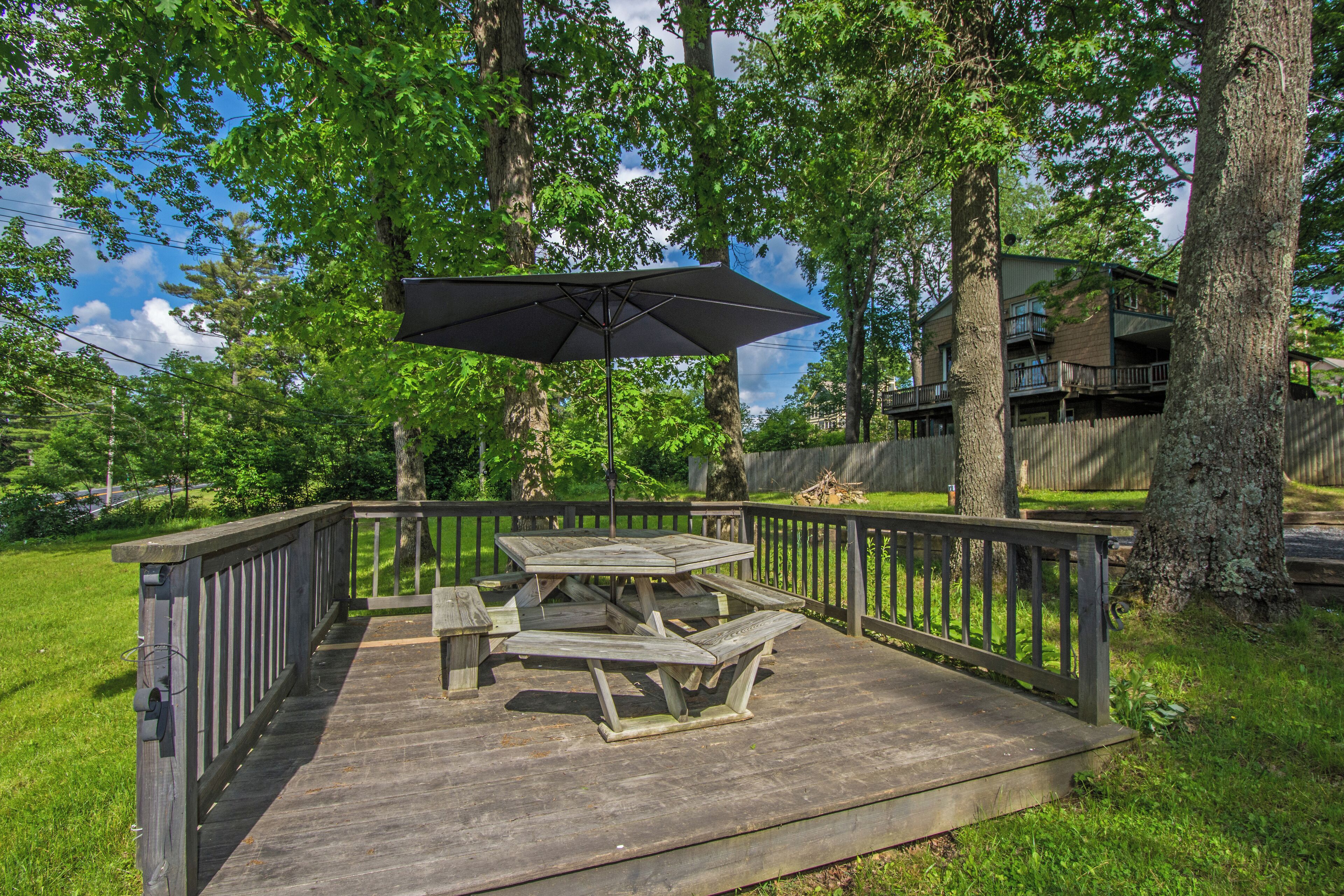 A wooden deck with picnic tables and an umbrella, surrounded by trees and a house in the background.
