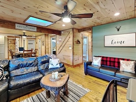 Un acogedor interior de cabaña con techo de madera, ventilador de techo, comedor con mesa y sillas, sala de estar con sofá azul y mesa de centro, y una pared con un ciervo montado y un letrero que dice "A GREAT PLACE TO HIBERNATE".