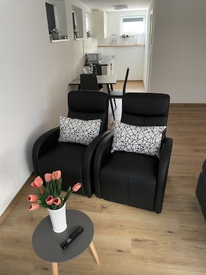Uma sala de estar moderna com duas poltronas pretas, uma mesa redonda pequena com um controle remoto e um vaso de tulipas, e uma área de jantar ao fundo.