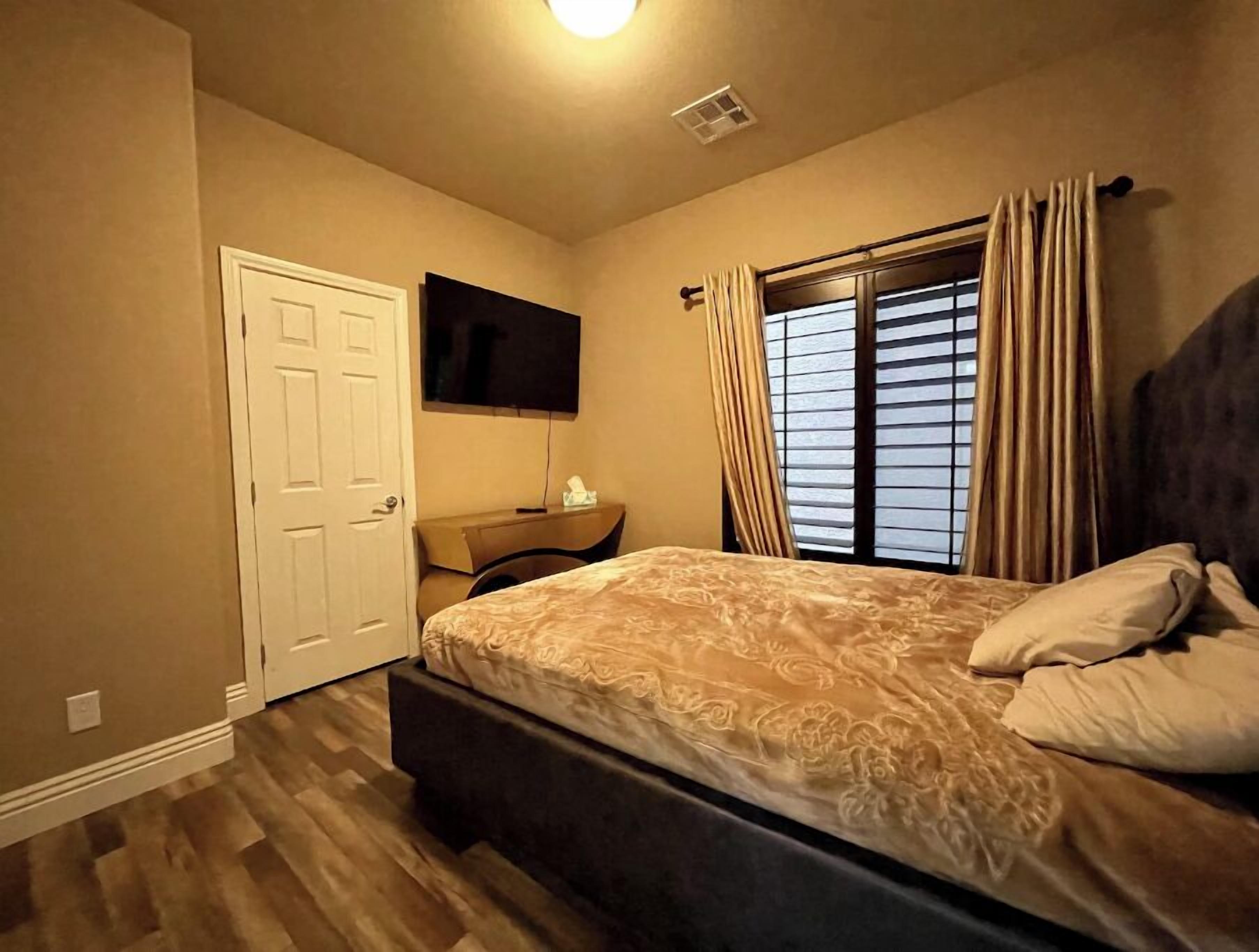A bedroom with a bed, a TV mounted on the wall, a desk, and a window with curtains.