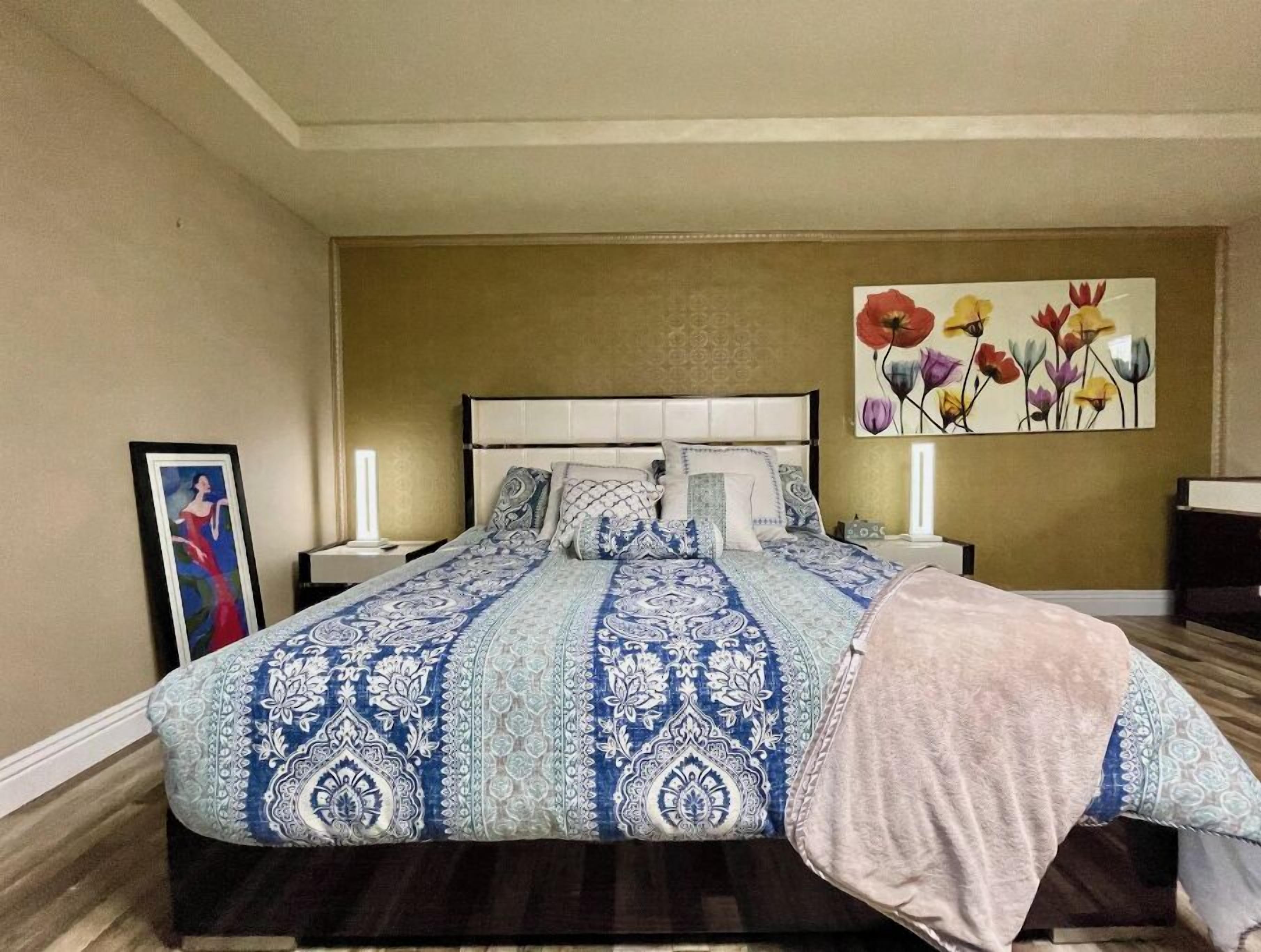 Un dormitorio con una cama grande, dos mesitas de noche, un cabecero y un cuadro con flores en la pared.