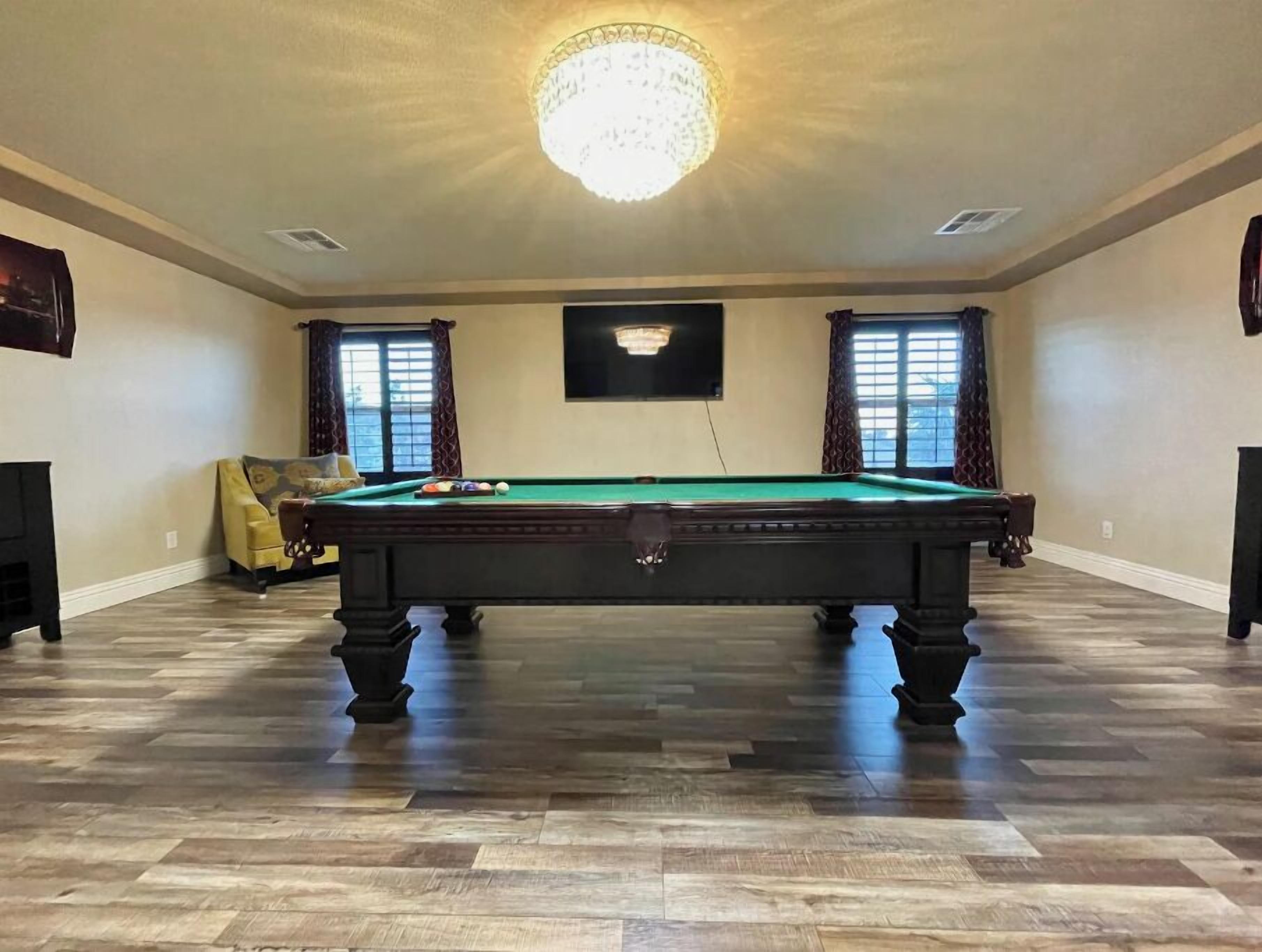 A spacious room with a pool table, two windows with curtains, a chandelier, and a flat-screen TV on the wall.
