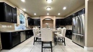 A modern kitchen with black cabinets, a stainless steel refrigerator, and a dining area with a marble countertop and upholstered chairs. - Luxury, Cheerful & Spacious 5 Bedroom Home! Free P (Las Vegas)