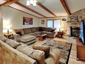 A cozy living room with a stone fireplace, wooden beams, and a large sectional sofa. - Knotty Pines by Taylor Made Deep Creek (Oakland)