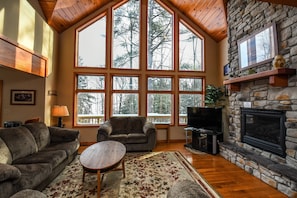 A cozy living room with a stone fireplace, large windows, a fireplace mantel, and a wooden ceiling. - Lake House by Taylor Made Deep Creek (Oakland)