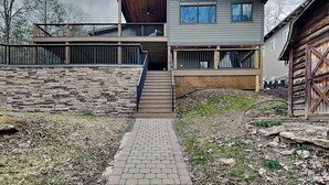 Una casa moderna de dos pisos con un muro de contención de piedra, una terraza de madera y un camino de ladrillo que conduce a la entrada.