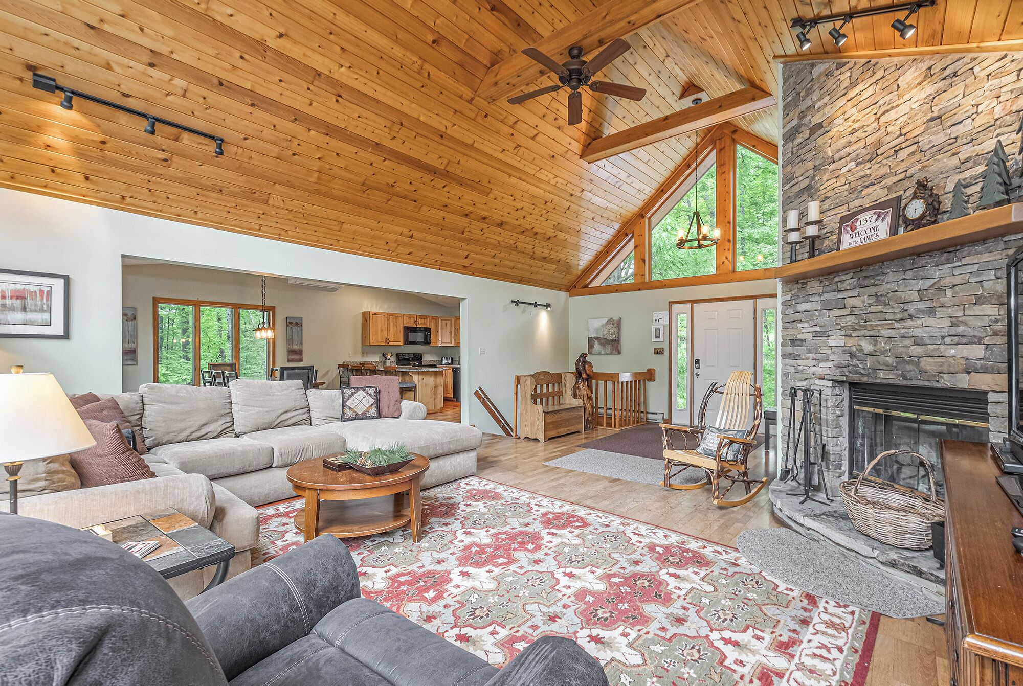 A spacious living room with a high ceiling, a stone fireplace, and a view of the kitchen and dining area.
