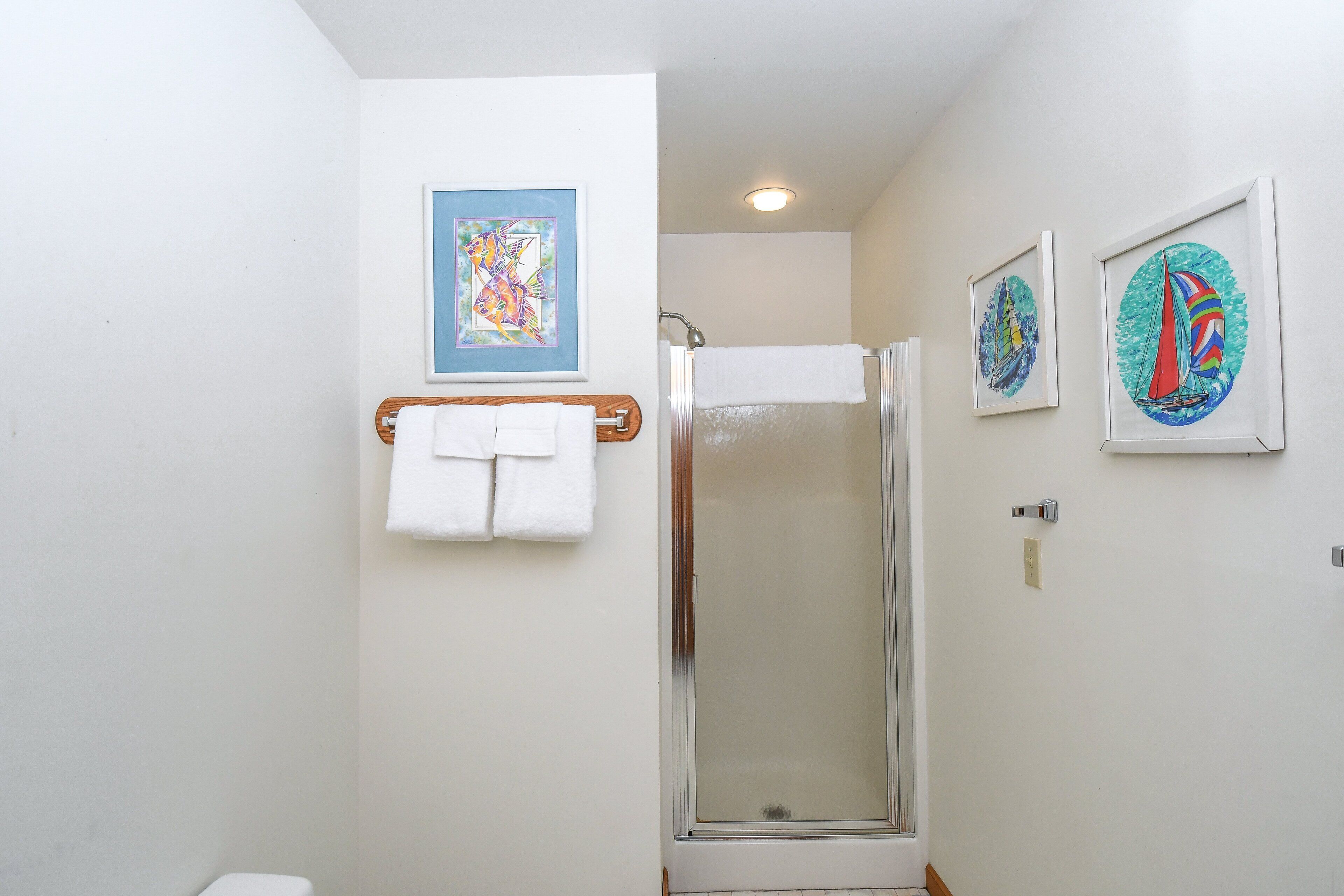 A bathroom with a shower, a towel rack with white towels, and two framed artworks on the wall.